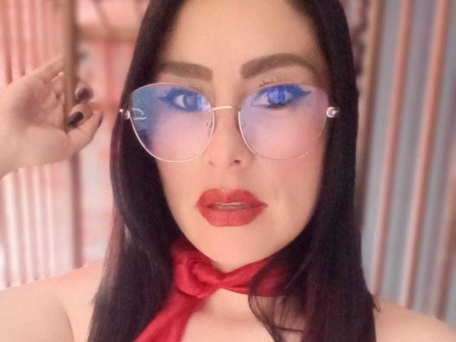 VioletaWhitee with blue-tinted aviator glasses, red lipstick, black hair, and a red scarf, holding her glasses with black-painted nails. Her eyes are strikingly blue.