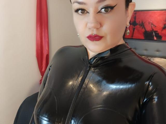 JadeLion, con cabello corto y oscuro, vestida con un brillante abrigo negro de látex con cremallera, tiene lápiz labial rojo, un piercing en la nariz y aretes, y mira a la cámara con una ligera sonrisa..