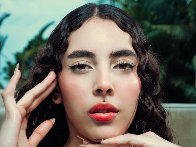 TianaVera has curly dark brown hair, a nose piercing, red glossy lips, and long eyelashes. Her hand is near her face, showing off her manicured nails.