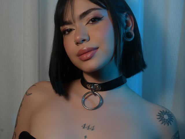 SalomeHil has black bob, nose ring, and large gauge earrings. She wears a black choker with a metal ring. Her tattoos include sun and lettering on her chest.
