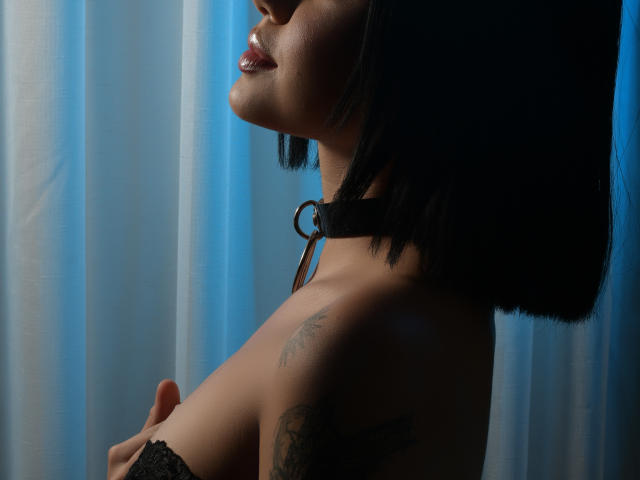 SalomeHil with dark bob, shiny lips, black lace top, covering her tits, showing a tattoo on her shoulder, against a blue curtain, with soft lighting.