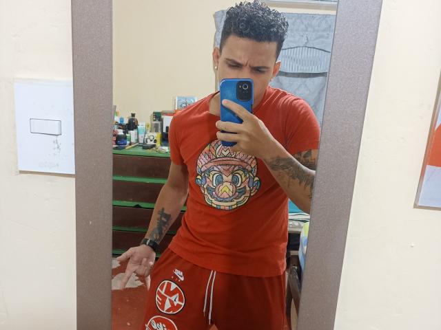 HottLoverGuy69 with curly black hair, tattooed arms, red t-shirt, and red shorts, taking a mirror selfie with a blue phone, looking annoyed, standing in a cluttered room.