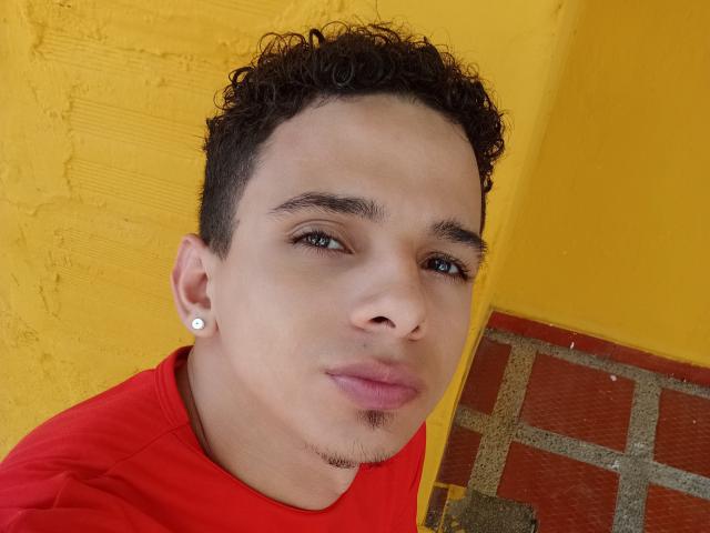HottLoverGuy69 has curly brown hair, light brown skin, green eyes, and a small goatee. He's wearing a red shirt, a white earring, and a slight pout.