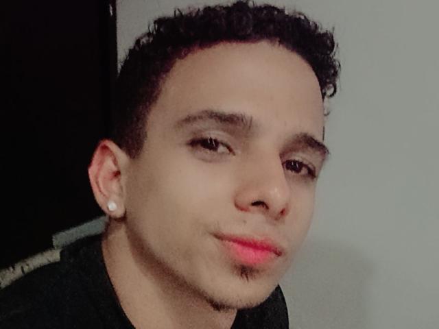 HottLoverGuy69 has curly dark hair, light brown skin, small nose, pink lips, slight goatee, and white ear studs, looking at camera with a slight smile.