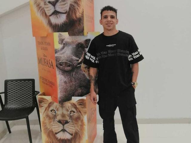 HottLoverGuy69 stands in front of a wall with three large lion portraits, wearing a black t-shirt with white text and black pants, looking happy.