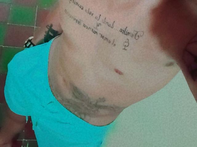 HottLoverGuy69's bare chest, showing tan lines, tattoos, and a small red mark, wearing bright blue shorts, with a visible hairy stomach and a black bracelet on his right wrist.