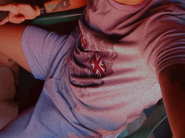 HottLoverGuy69's shirt has a Union Jack print, wearing a purple-gray tee, showing a tattooed arm, and making a peace sign. His ass is slightly visible.