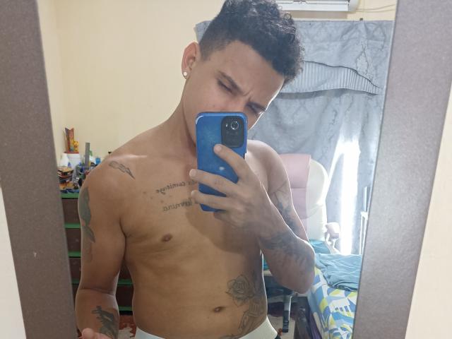 Shirtless HottLoverGuy69 with short curly hair, ear piercings, tattoos, holding blue phone, mirror selfie, visible chest and abdominal tattoos, white underwear, looking slightly annoyed.