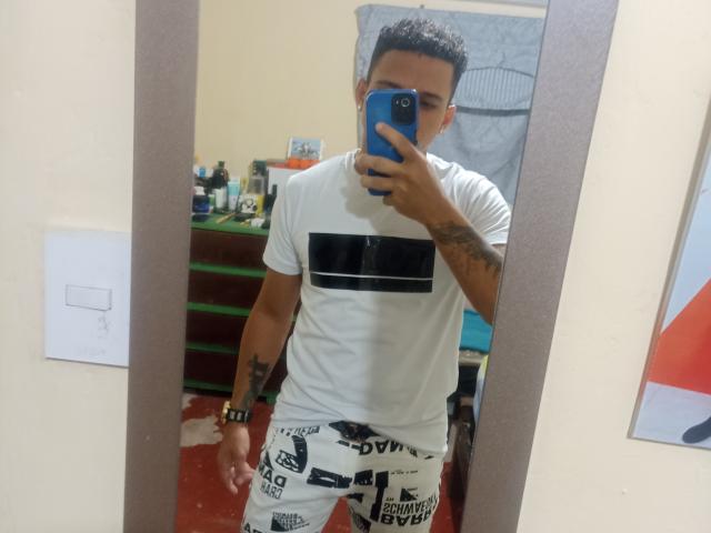 HottLoverGuy69 with short dark hair, tattoos on arms, wearing white t-shirt with black stripe and white pants with black text, holding blue phone in mirror selfie.