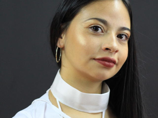 ZuriLaoni tem olhos castanhos, pele clara, cabelo preto e liso, e usa um colar branco com brincos de argola, olhando ligeiramente para o lado com um sorriso discreto..