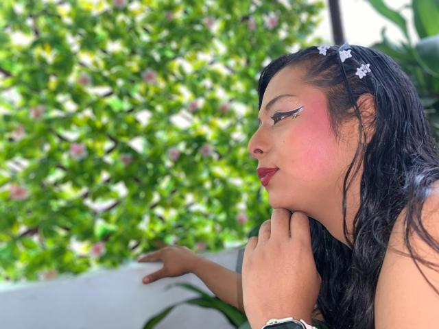 EmilyTsSweet with wet black hair, pink blushed cheeks, winged eyeliner, red lipstick, and white hair clips, resting her chin on her hand, looking to the side, outdoors.