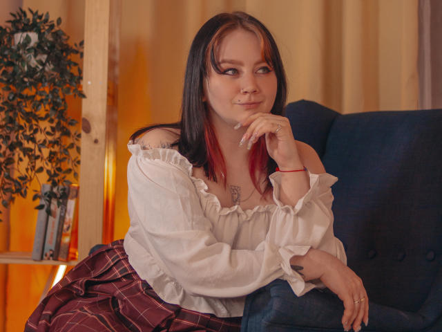 LunaLise with dark brown and red-tipped hair, off-shoulder white blouse, and red plaid skirt, sitting on a blue chair, hand near mouth, smiling slightly.