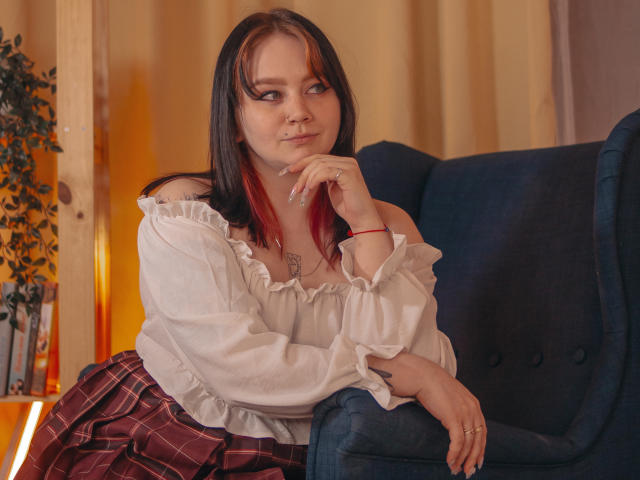 LunaLise with red and black hair, white off-shoulder blouse, plaid skirt, and red bracelet, sitting in a blue chair, resting her chin on her hand, looking to the side.