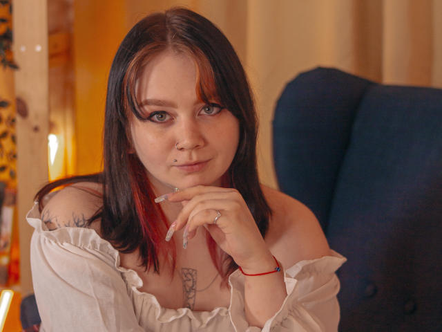 LunaLise with shoulder tattoos, red-black hair, off-shoulder white top, ring on finger, nose piercing, subtle smile, hand on chin, sitting in blue chair, looking at camera.