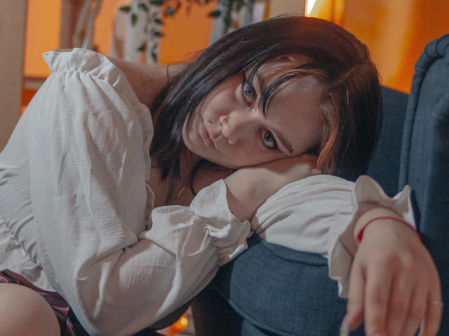 LunaLise with brown and orange-streaked hair, nose ring, white off-shoulder shirt, leaning on blue chair, looking at camera with a pouty expression, red bracelet on right wrist.