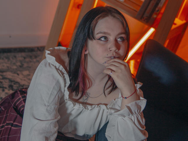 LunaLise with pale skin, brown and pink dyed hair, white off-shoulder top, and plaid skirt, pouting with finger to mouth, looking to the side, in dim lighting.