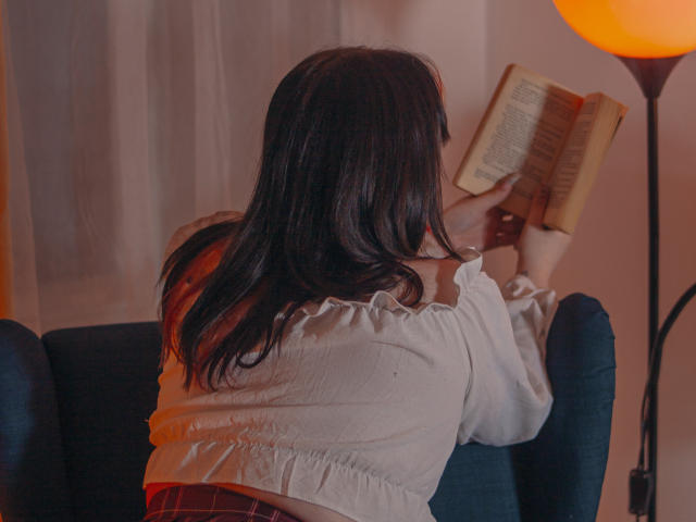 LunaLise with long dark hair, wearing a white off-shoulder top and red plaid skirt, reading a book on a blue armchair, holding it with both hands.