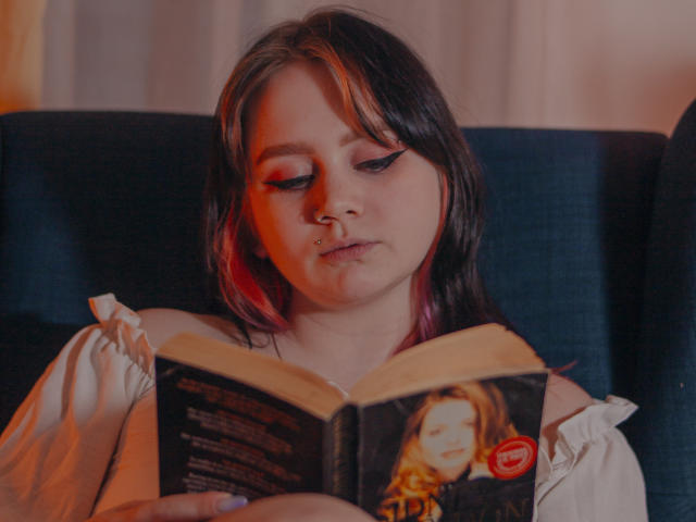 LunaLise with pink and black hair, nose ring, and winged eyeliner, reading a book with a cover featuring a woman's face. She's in a blue chair, wearing a white top.