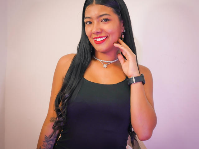 Ailyn Campos, con lunghi capelli neri, rossetto rosso, canotta nera, collana, orologio al polso, e che sorride alla fotocamera, mostrando un tatuaggio sul braccio destro. La sua mano tocca la guancia..
