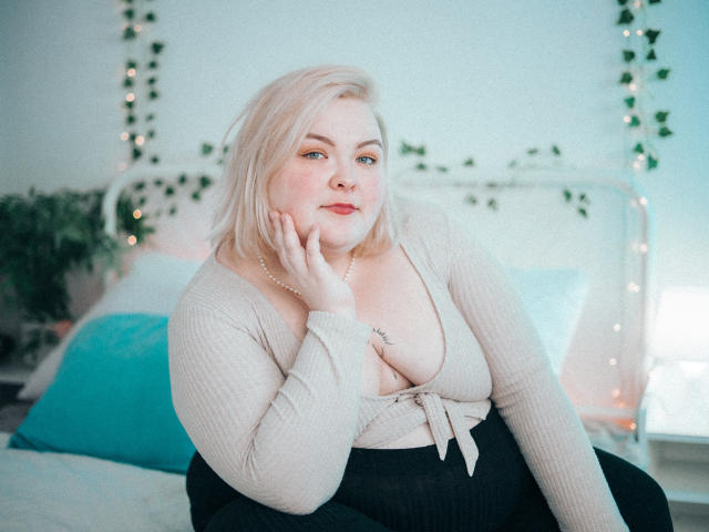 EllaBigBabe met blond haar, die een beige geribbelde top draagt, waarbij haar grote borsten zichtbaar zijn, zwarte broek, die haar wang aanraakt, zittend op een bed met turquoise kussens..