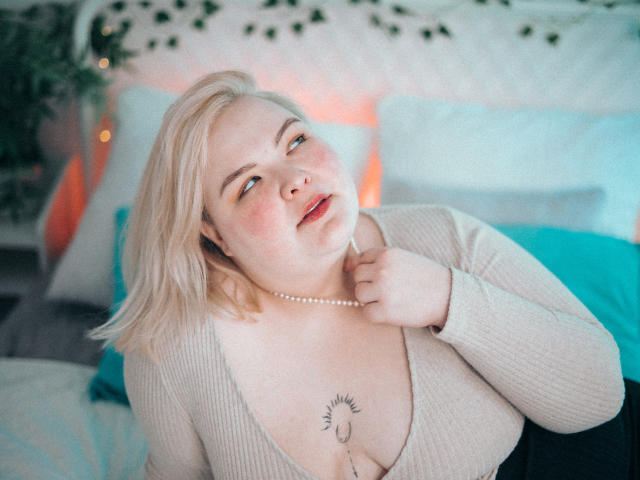 EllaBigBabe with blonde hair, light skin, and a tattoo under her tits, wearing a pearl necklace, looks up with a slight smile, hand near her neck.