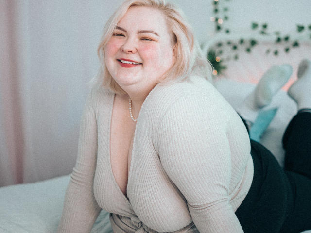 EllaBigBabe with blonde hair, smiling, wearing a deep V-neck beige ribbed top, showing her big tits, black pants, and a pearl necklace, sitting on a bed.