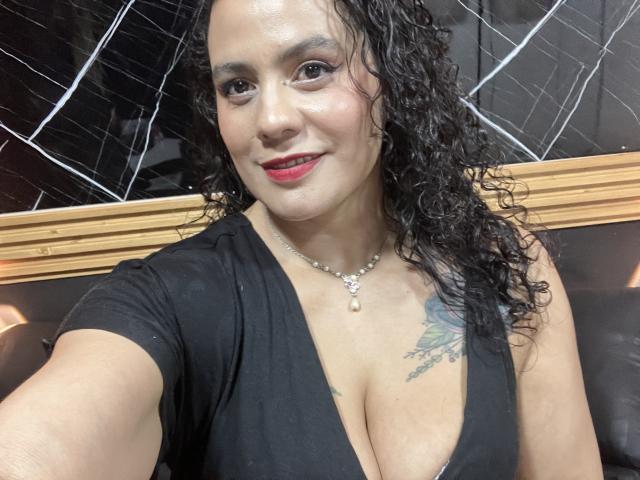 Tamarajoones, con capelli neri ricci, rossetto rosso e un abito nero che mette in mostra un seno prosperoso, indossa una collana, ha un tatuaggio sul petto destro, sorride ed è seduta in una stanza buia..