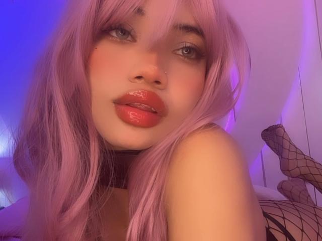 LiliMays with pink hair, shiny red lips, black fishnet, and a nose ring, looking at the camera with blue and purple lighting. Her tits are slightly visible.