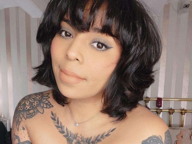 Chili, with black shoulder-length hair and bangs, has brown eyes and a smug expression, showing her tattooed shoulders and chest.