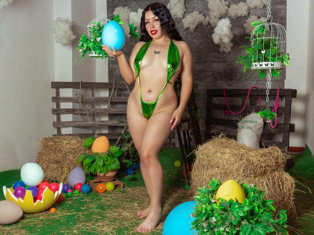 KateWelsin with dark curly hair, green micro bikini, holding blue egg, standing on hay, surrounded by eggs and green plants, showing big ass and small tits, red lipstick.