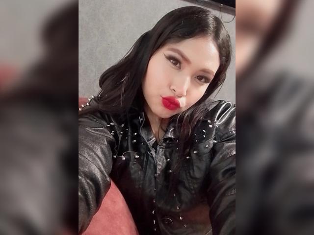 OliviGrace, con lunghi capelli neri, rossetto rosso e una giacca di pelle nera con borchie argentate, guarda la fotocamera con un leggero sbuffo. Primo piano sfocato..