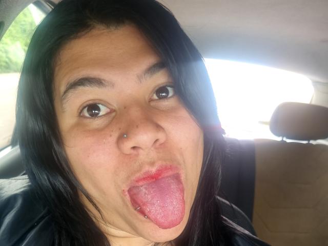 Das Gesicht von AlmaWhite69 in einem Auto, mit schwarzem Haar, braunen Augen, einem Nasenpiercing und der Zunge herausgestreckt, wobei ein Zungenspiercing zu sehen ist. Sie schaut in die Kamera und hat die Zunge heraus..