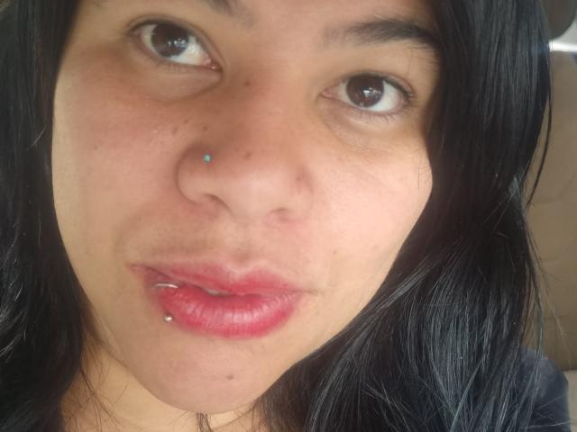 AlmaWhite69 hat ein Nasenpiercing und ein Lippenpiercing, hat dunkles Haar, braune Augen und rote Lippen. Sie blickt nach oben mit leicht geöffnetem, glänzend rosa Mund..