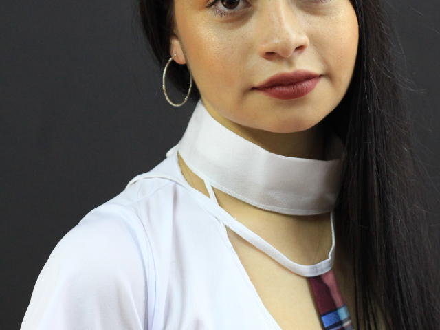 ZuriLaoni ha una sciarpa bianca al collo, orecchini a cerchio, occhi marroni e lunghi capelli neri, indossa una maglietta bianca e una cravatta a scacchi rossi, e guarda leggermente verso destra..
