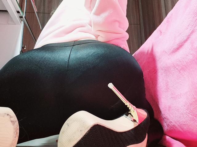 A traseira preta e brilhante de Akkanee está voltada para a câmera, vestindo calças pretas de cintura alta e justas, e um moletom rosa. Ela está usando um salto alto branco com uma tira vermelha..