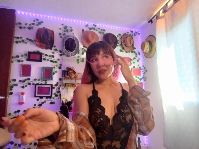 SugarMuse69 with pink hair, wearing black lace lingerie, plaid shirt off-shoulder, holding orange nail polish, winking, purple LED lights in background, hats and plants on wall.