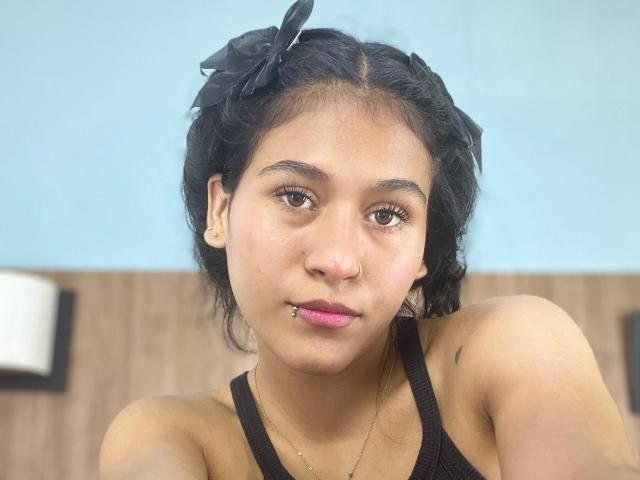 AnnieDarling with dark curly hair, black bow, nose ring, and lip piercing, wearing a black tank top, looking at the camera with a neutral expression.