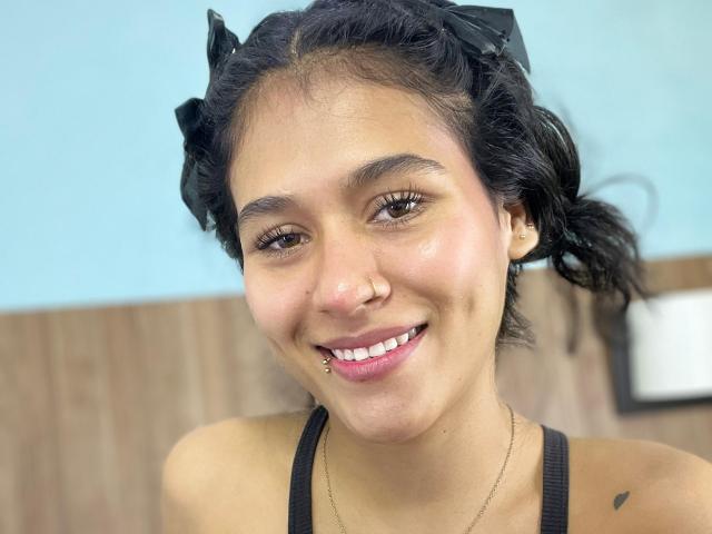 AnnieDarling has brown eyes, black curly hair with black ribbons, a nose ring, a lip piercing, and a small heart tattoo on her right shoulder. She's smiling.