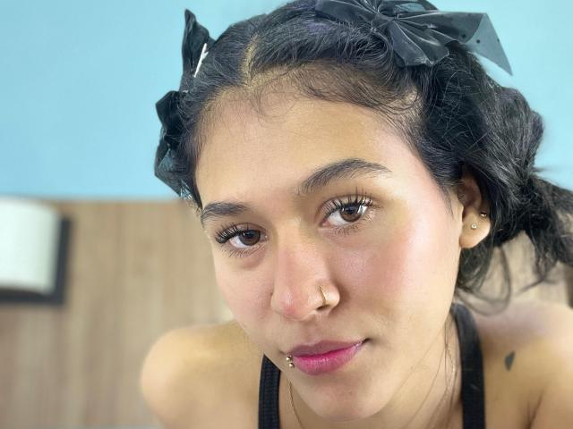 AnnieDarling has dark hair in black bows, brown eyes, pink lipstick, nose ring, and lip ring, with a small tattoo on her shoulder. She's wearing a black top.