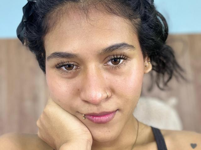 AnnieDarling has brown eyes, a nose ring, and a lip piercing. She's resting her chin on her hand with curly black hair, a slight smile, and pink lipstick.