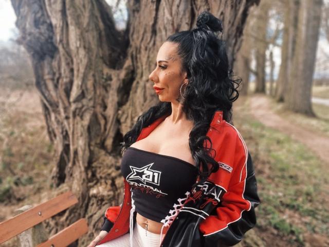 CandySuck: una ragazza con capelli neri raccolti in uno chignon, indossa una giacca rossa e un top nero a spalline che mette in mostra un seno generoso, è in piedi vicino a un albero, guarda di lato, con rossetto rosso..