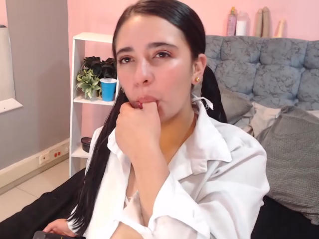 Laila Rogerts, com maria-chiquinhas pretas, camisa branca e pele rosada, chupando o dedo, olhando para a câmera, com uma expressão fofa e sexy, sentada em uma cama preta..