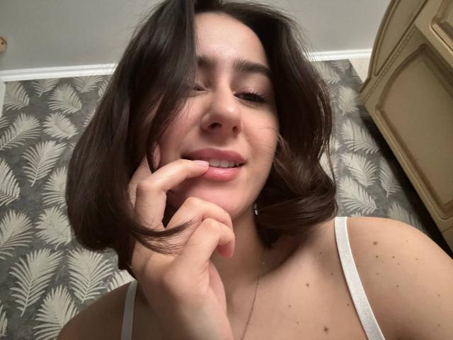 KamilaHarison with brown wavy hair, white tank top, smiling, eyes half-closed, biting finger, showing small nose, subtle makeup, cute, with a small mole on her right cheek.