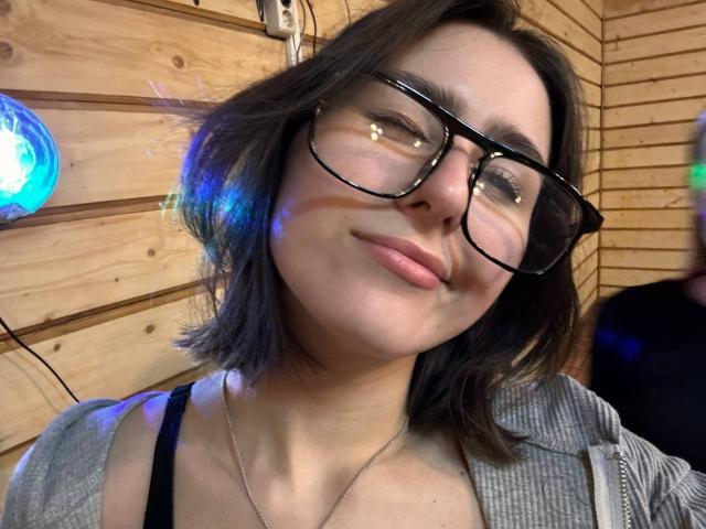 KamilaHarison with glasses, short brown hair, light skin, subtle smile, wearing a gray top and black strap, looking slightly up at the camera. Boobs visible under the top.