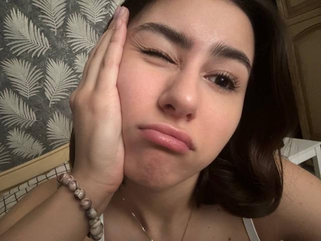 KamilaHarison with one eye closed, winking, touching her face with one hand, wearing a white tank top, and a bead bracelet on her wrist, has slightly puffy lips.
