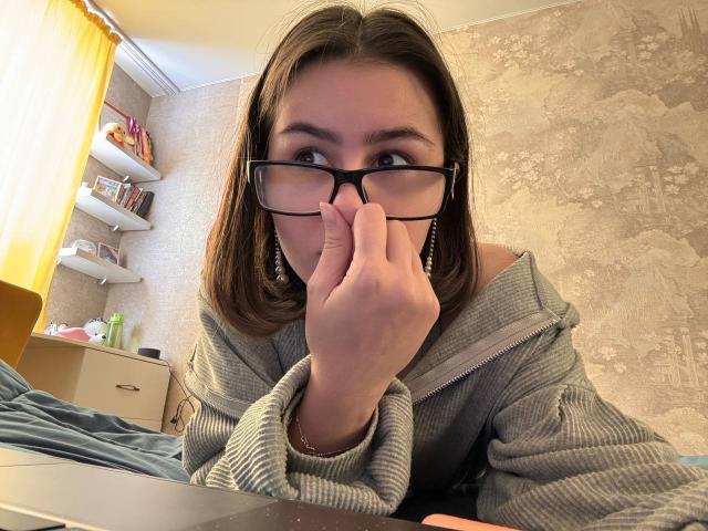 KamilaHarison with glasses, gray hoodie, and earrings, biting her finger, looking at camera, brown hair, laptop in front, yellow curtain, bookshelf, and wall in background.