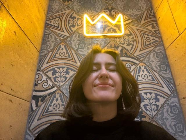 KamilaHarison with her eyes closed, smiling, wearing a black top, has medium-length brown hair, and a neon crown light above her head. Her nose is slightly upturned.