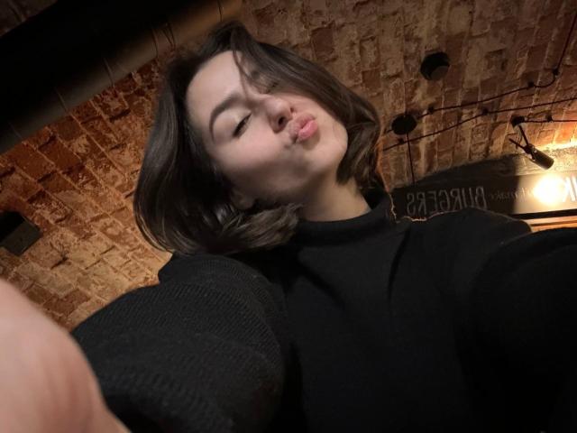 KamilaHarison with pouty lips, black sweater, short brown hair, slightly tilted head, and a selfie angle, in a dimly lit brick ceiling room.