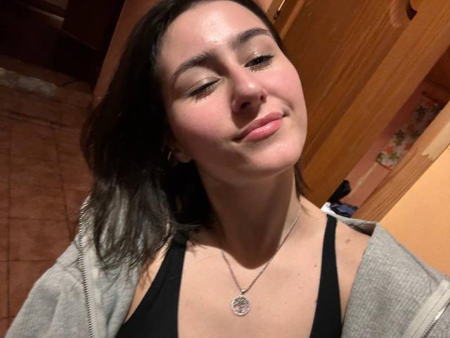KamilaHarison with closed eyes, slight smile, wearing a black tank top, gray hoodie, and silver necklace with a circular pendant, has medium-length dark brown hair.