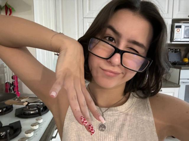 KamilaHarison with winking eye, black glasses, red nail polish, gray sleeveless top, necklace, and raised arm in kitchen. Boobless, cute, casual, smiling.
